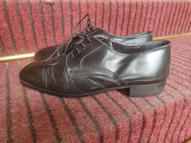 May include: A pair of black leather dress shoes with laces. The shoes have a classic design with a rounded toe and a low heel. The shoes are sitting on a red carpet.
