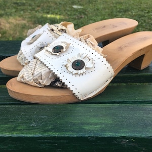 Puede incluir: Zuecos de cuero blanco con tacón de madera y adornos de flores verdes. Los zuecos tienen un diseño recortado y están decorados con trozos de periódico antiguo.