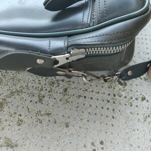 May include: A dark green leather bag with a zipper closure and a metal hook. The bag has a strap with a metal buckle.