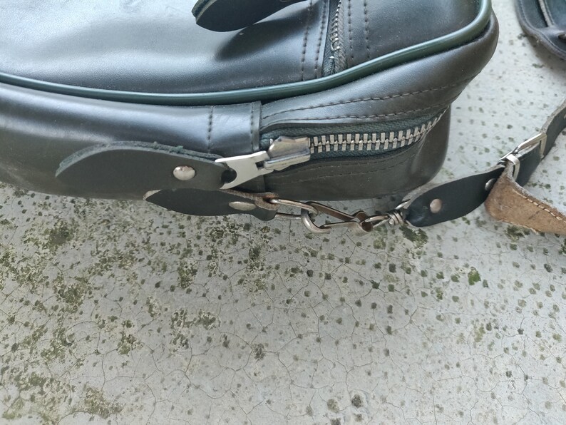 May include: A dark green leather bag with a zipper closure and a metal hook. The bag has a strap with a metal buckle.