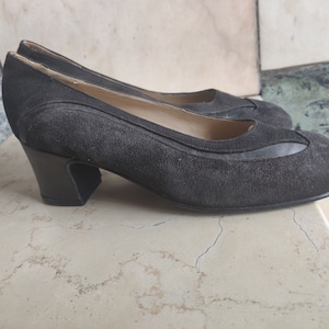 Puede incluir: Un par de zapatos de tacón de ante gris oscuro con tacón negro. Los zapatos tienen un diseño sencillo con punta redonda.