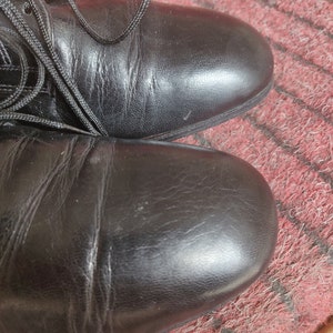 May include: Close-up of two black leather boots with laces. The boots are on a red and black patterned surface. The leather has visible creases and a slight sheen, suggesting regular wear.