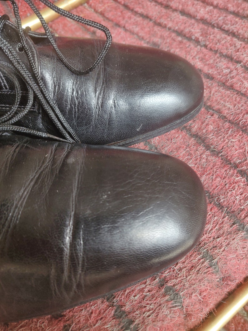 May include: Close-up of two black leather boots with laces. The boots are on a red and black patterned surface. The leather has visible creases and a slight sheen, suggesting regular wear.