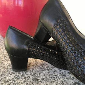Puede incluir: Un par de zapatos de tac&oacute;n de cuero negro con un patr&oacute;n tejido. Los zapatos tienen un tac&oacute;n de bloque bajo y una punta cerrada.