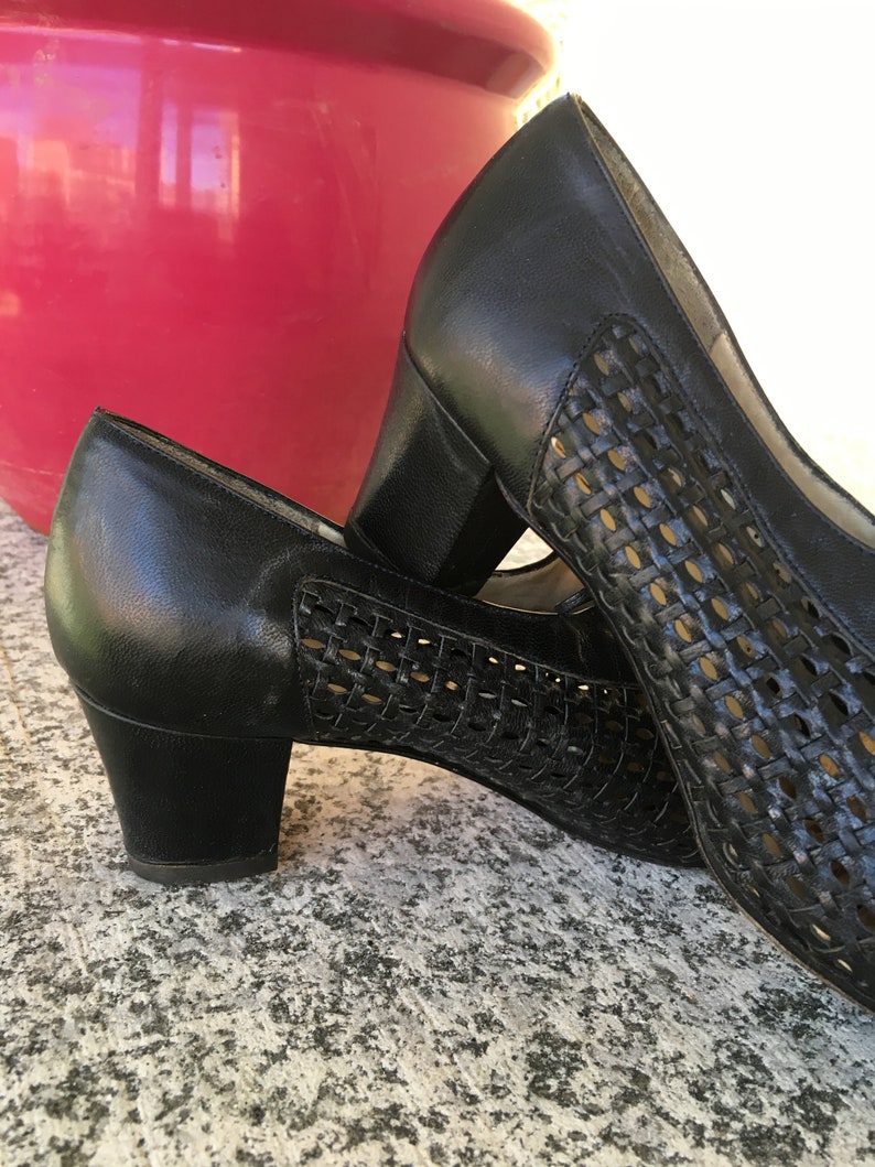 Puede incluir: Un par de zapatos de tac&oacute;n de cuero negro con un patr&oacute;n tejido. Los zapatos tienen un tac&oacute;n de bloque bajo y una punta cerrada.