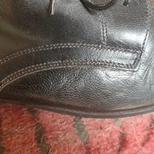 May include: Close-up of a black leather shoe, showcasing the textured surface and stitching details. The laces are visible at the top. The shoe appears to be new or well-maintained, with a focus on the craftsmanship.