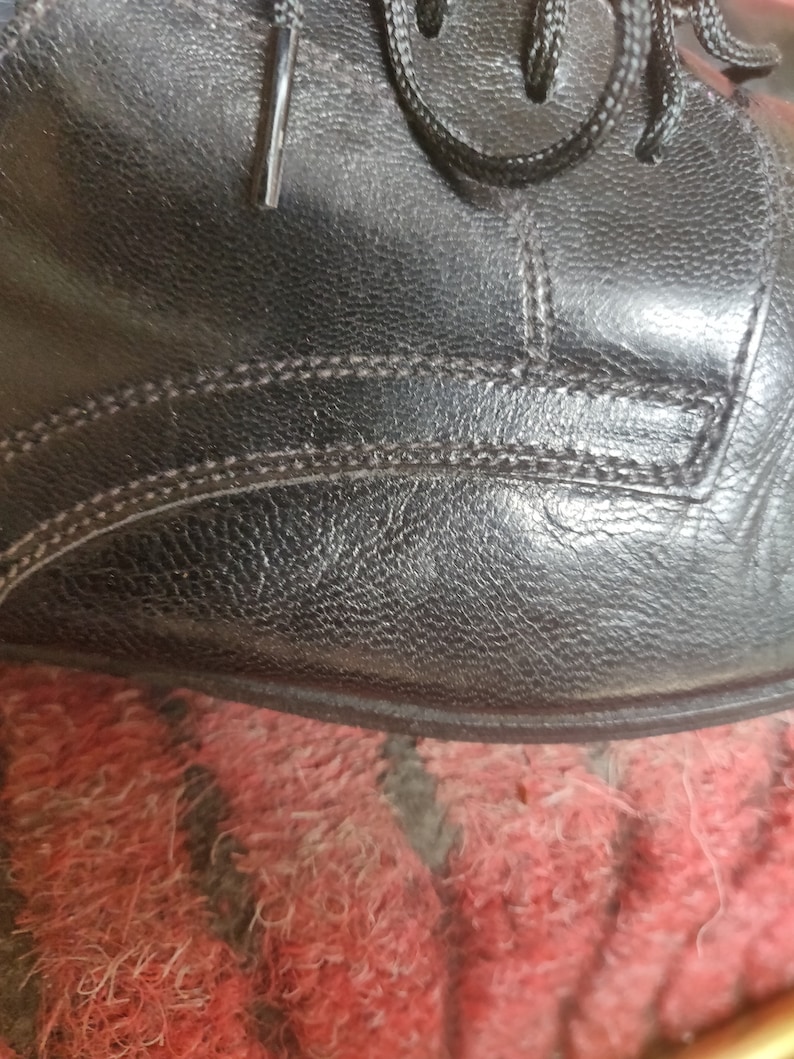 May include: Close-up of a black leather shoe, showcasing the textured surface and stitching details. The laces are visible at the top. The shoe appears to be new or well-maintained, with a focus on the craftsmanship.