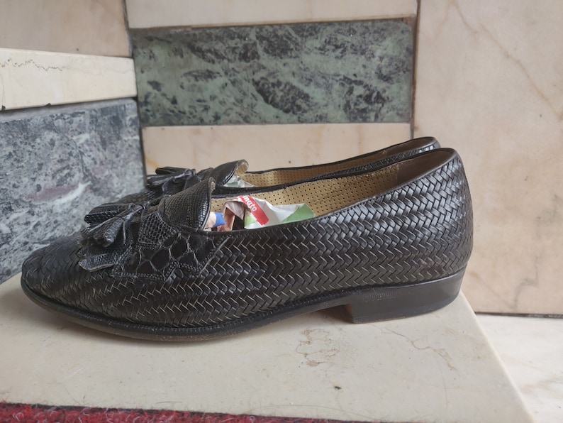 K&ouml;nnte beinhalten: Schwarze Leder-Tasssel-Slipper mit gewebtem Muster. Die Slipper haben eine spitze Zehenpartie und einen niedrigen Absatz.