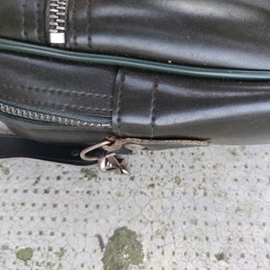May include: Close-up of a dark green leather bag with a zipper and a metal clasp. The bag has a green trim around the edge.