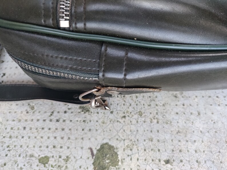 May include: Close-up of a dark green leather bag with a zipper and a metal clasp. The bag has a green trim around the edge.