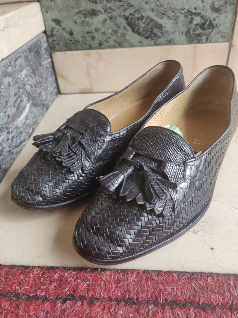 K&ouml;nnte beinhalten: Ein Paar schwarze Leder-Tasssel-Slipper mit einem gewebten Muster. Die Slipper haben eine spitze Zehenpartie und einen niedrigen Absatz.