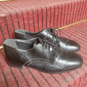 May include: A pair of black leather dress shoes with laces, resting on a red carpeted staircase. The shoes have a classic design with visible stitching and a polished finish, suitable for formal occasions.