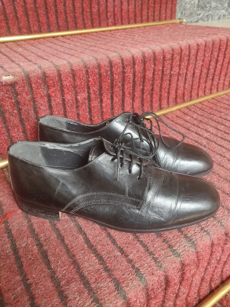 May include: A pair of black leather dress shoes with laces, resting on a red carpeted staircase. The shoes have a classic design with visible stitching and a polished finish, suitable for formal occasions.