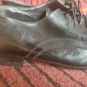May include: Close-up of a pair of black leather dress shoes. The shoes have a classic Oxford design with laces and visible stitching. The leather shows signs of wear, adding character to the footwear. The shoes are on a red and brown textured surface.
