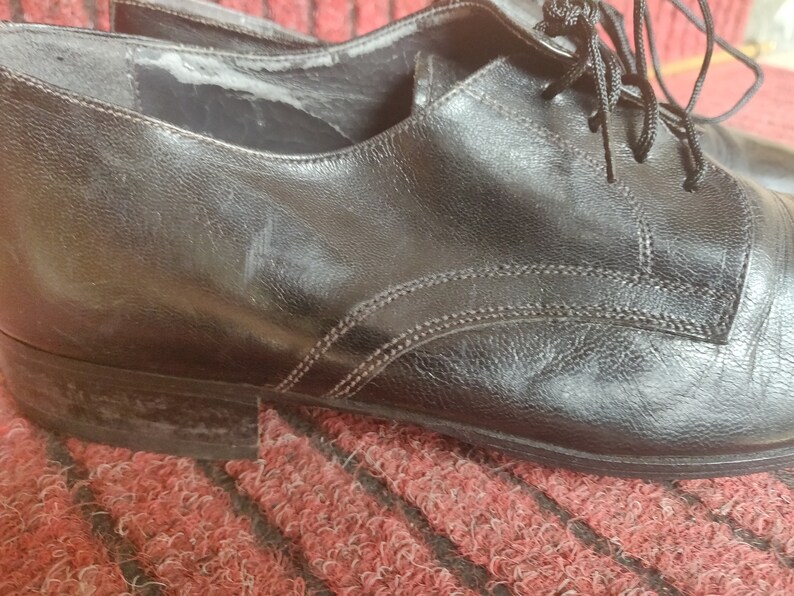 May include: Close-up of a pair of black leather dress shoes. The shoes have a classic Oxford design with laces and visible stitching. The leather shows signs of wear, adding character to the footwear. The shoes are on a red and brown textured surface.