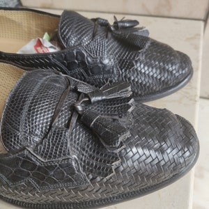K&ouml;nnte beinhalten: Ein Paar schwarze Leder-Tasssel-Slipper mit einem gewebten Muster. Die Slipper haben eine spitze Zehenpartie und einen niedrigen Absatz.