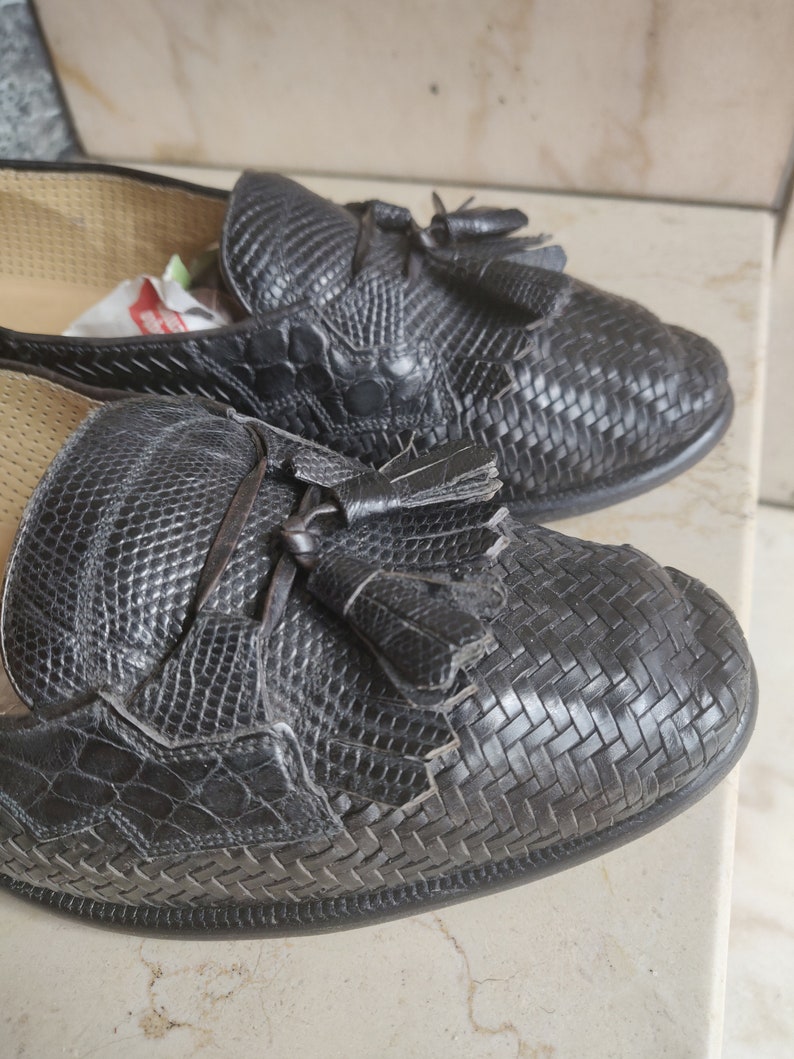 K&ouml;nnte beinhalten: Ein Paar schwarze Leder-Tasssel-Slipper mit einem gewebten Muster. Die Slipper haben eine spitze Zehenpartie und einen niedrigen Absatz.