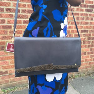 Op de afbeelding: Een donkerblauwe leren schoudertas met een slangenleren rand. De tas heeft een gouden sluiting en een lange band. De tas wordt gedragen door iemand die een blauwe en zwarte bloemenjurk draagt.