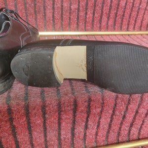 May include: Black leather dress shoes with a rubber sole. The sole is textured with a ribbed pattern. The heel is made of a lighter-colored material. The shoes are on a red and brown patterned surface.