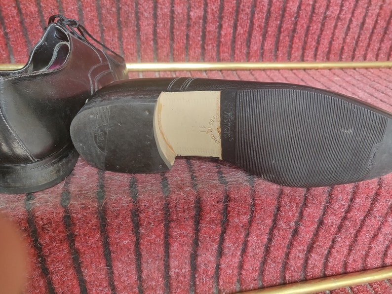 May include: Black leather dress shoes with a rubber sole. The sole is textured with a ribbed pattern. The heel is made of a lighter-colored material. The shoes are on a red and brown patterned surface.