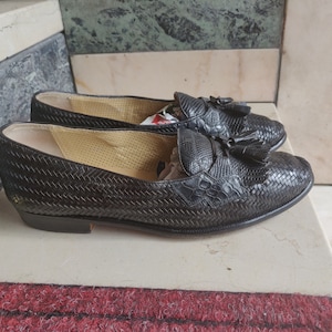 K&ouml;nnte beinhalten: Ein Paar schwarze Leder-Slipper mit Quasten. Die Slipper haben ein gewebtes Muster und sind aus einem strukturierten Material gefertigt.
