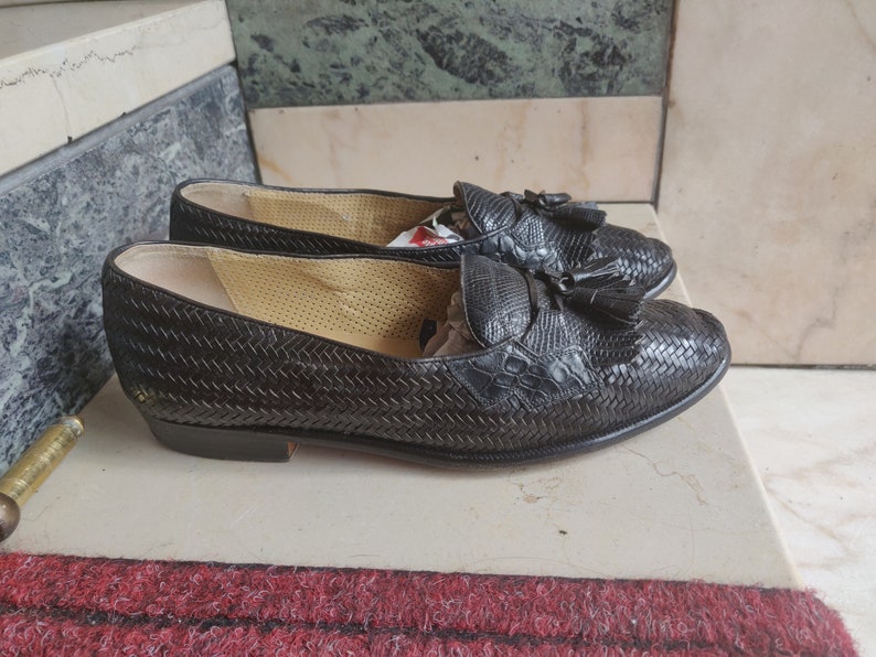 K&ouml;nnte beinhalten: Ein Paar schwarze Leder-Slipper mit Quasten. Die Slipper haben ein gewebtes Muster und sind aus einem strukturierten Material gefertigt.