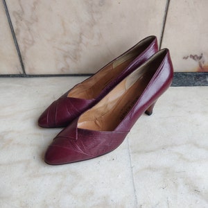 Può includere: Un paio di scarpe con tacco in pelle bordeaux con punta a punta. Le scarpe hanno un design in pelle testurizzata e il nome del marchio "Michelangelo" è visibile all'interno della scarpa.