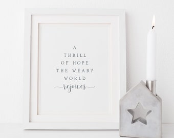 May include: A white framed print with the text "A thrill of hope the weary world rejoices". The print is sitting on a white surface next to a white candle in a gray concrete house-shaped candle holder.