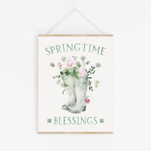 以下が含まれることがあります： 「SPRINGTIME BLESSINGS」と緑色の文字が書かれた白い看板。看板には、ピンクの花と緑の葉がついた緑色のレインブーツ2つが描かれた水彩画が描かれている。