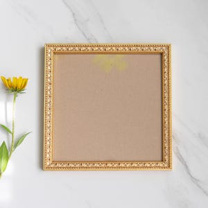 Könnte beinhalten: Ein goldfarbener, verzierter Bilderrahmen mit braunem Innenraum liegt auf einer weißen Marmoroberfläche. Eine gelbe Blume mit grünen Blättern befindet sich links vom Rahmen. Der Rahmen hat einen detaillierten, strukturierten Rand.