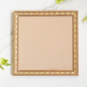 May include: Square gold picture frame with decorative detailing. The frame has a light brown backing and is set against a white marble surface. Green foliage accents the scene.