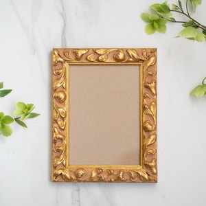 May include: A gold-coloured picture frame with an ornate design. The frame has a brown mount and is empty.