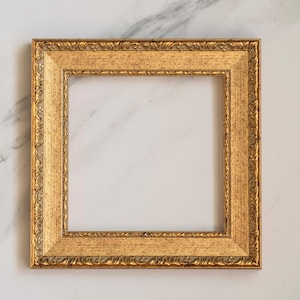 Pode incluir: Uma moldura quadrada com acabamento dourado metálico. A moldura apresenta um padrão ornamental em relevo semelhante a folhas ao longo das bordas externas e internas, rodeando uma borda central lisa e plana.