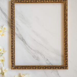 May include: A rectangular picture frame with an ornate, textured gold finish featuring repeating leaf and oval patterns along the border. The frame is empty, showcasing a hollow centre, and is positioned vertically with a metallic gold sheen.