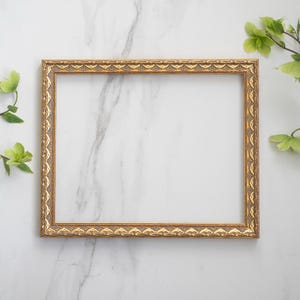 May include: A gold picture frame with an ornate design sits on a white marble surface. Green leaves are on either side of the frame.