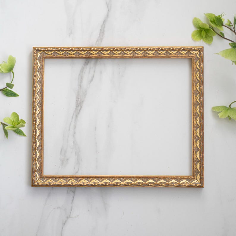 May include: A gold picture frame with an ornate design sits on a white marble surface. Green leaves are on either side of the frame.