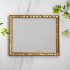 May include: A gold-colored picture frame with an ornate design sits on a white marble surface. The frame is empty and ready for a photo or artwork.