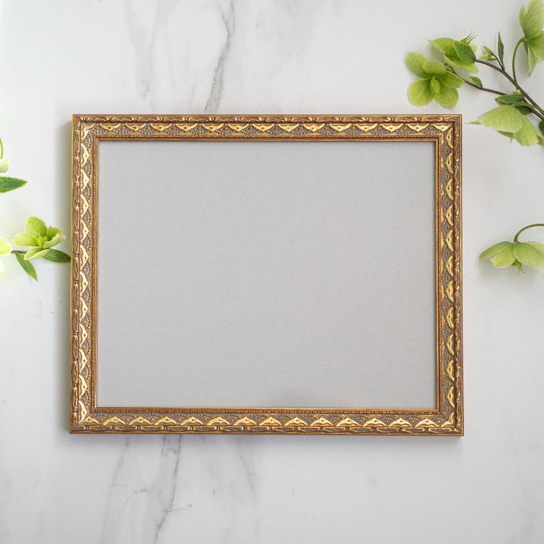 May include: A gold-colored picture frame with an ornate design sits on a white marble surface. The frame is empty and ready for a photo or artwork.
