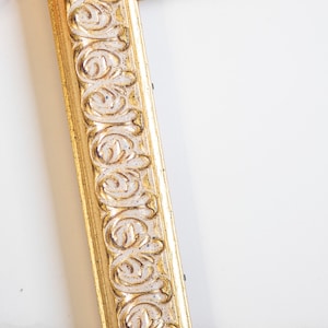 May include: A close-up of a gold picture frame with an ornate design. The frame is made of wood and has a white background.
