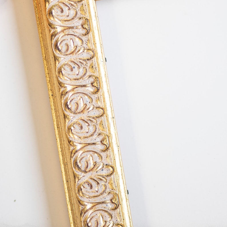 May include: A close-up of a gold picture frame with an ornate design. The frame is made of wood and has a white background.