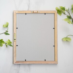 May include: A light wood picture frame with a white mat and black mounting clips. The frame is on a white marble surface.