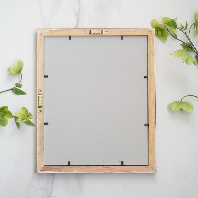 May include: A light wood picture frame with a white mat and black mounting clips. The frame is on a white marble surface.