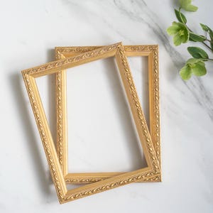Op de afbeelding: Twee rechthoekige fotolijsten met een metallic gouden afwerking en een sierlijk, verhoogd bladvormig patroon langs de binnen- en buitenranden. De lijsten zijn in een lichte hoek geplaatst, overlappen elkaar en tonen een gedetailleerde textuur.