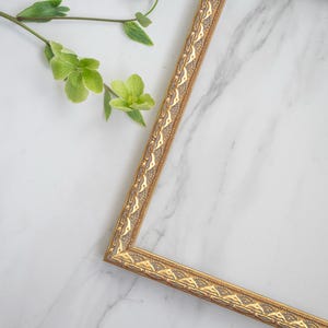 May include: A gold picture frame with an ornate design sits on a white marble surface. Green leaves and flowers are in the upper left corner of the image.