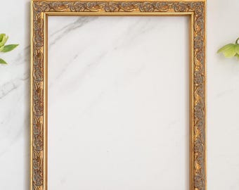 May include: A rectangular picture frame featuring a metallic gold finish with intricate, raised floral and scrollwork detailing. The inner and outer edges of the frame are smooth and gold, while the recessed center band has a dark, textured finish.