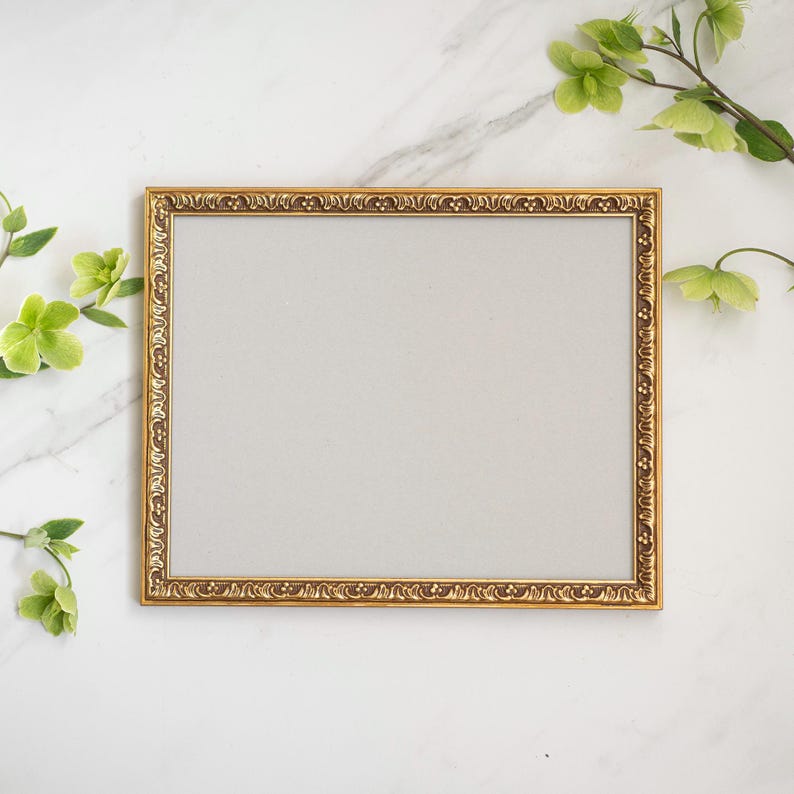 May include: A gold picture frame with an ornate design sits on a white marble surface. Green leaves and flowers are arranged around the frame.
