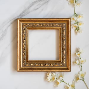 Könnte beinhalten: Ein quadratischer Bilderrahmen mit metallischem Goldfinish und einem verzierten, erhabenen Schnörkelmuster entlang des Innenrandes. Der Rahmen hat einen glatten, mattgrauen vertieften Bereich zwischen dem Außenrand und der Zierleiste.