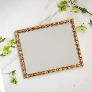 May include: A gold-colored picture frame with an ornate design. The frame is on a white marble surface with green leaves.