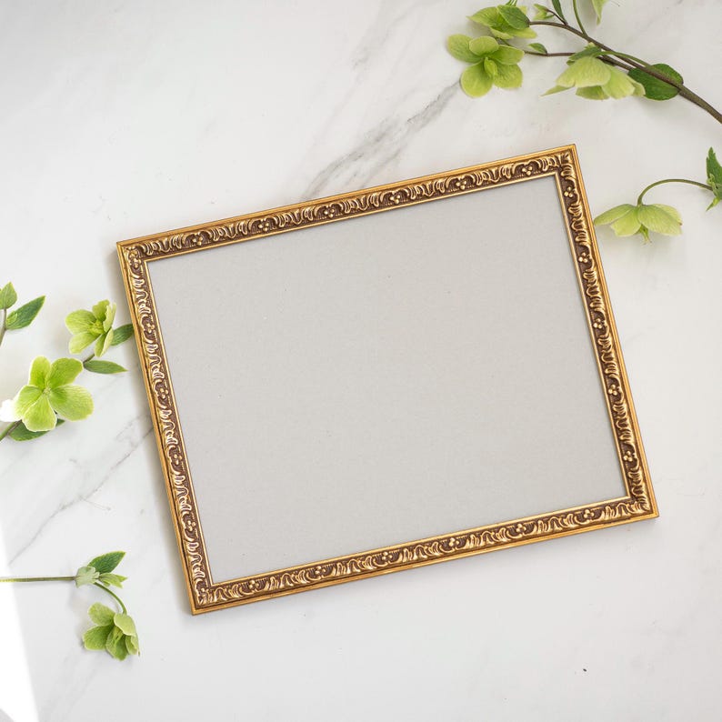 May include: A gold-colored picture frame with an ornate design. The frame is on a white marble surface with green leaves.