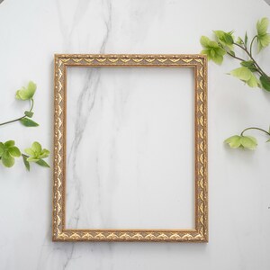 May include: A gold picture frame with an ornate design sits on a white marble surface. Green leaves and flowers are arranged around the frame.